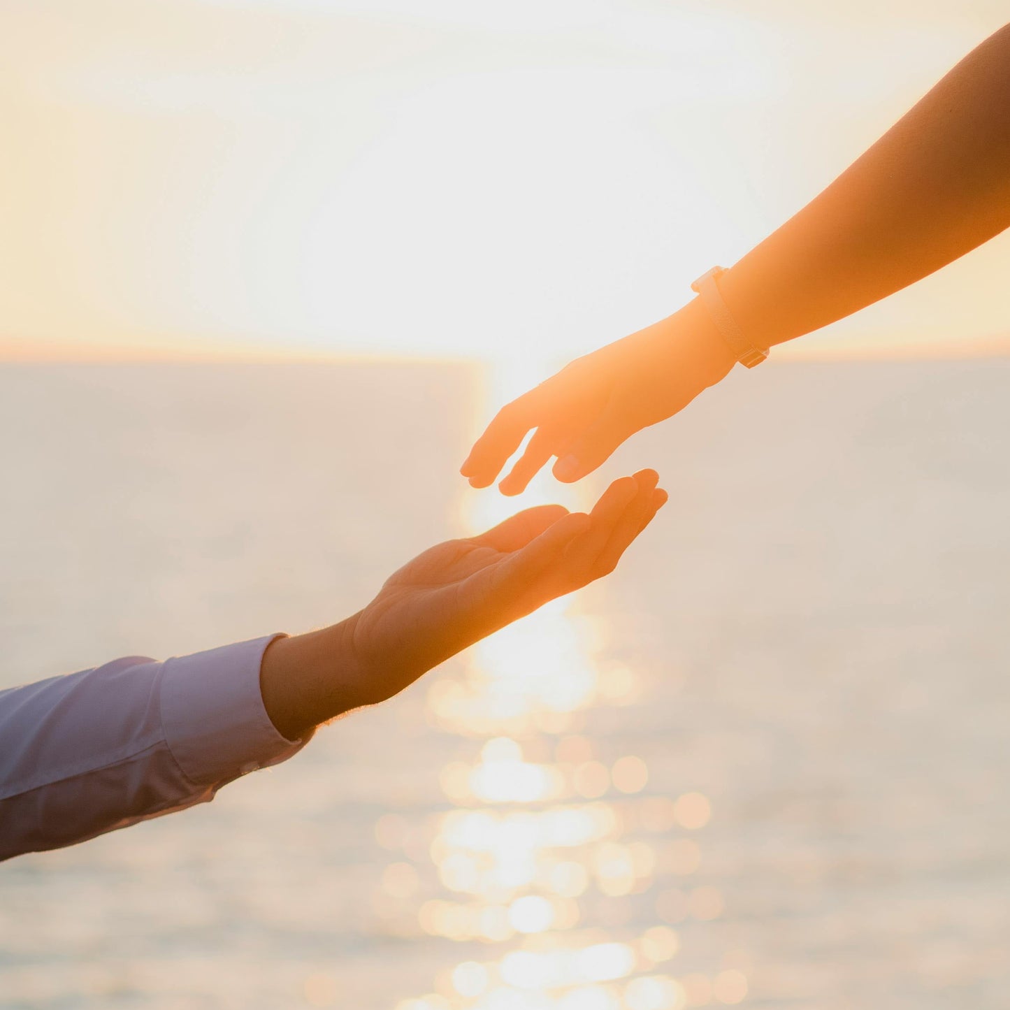 Two hands reaching towards each other over a sunset over water.